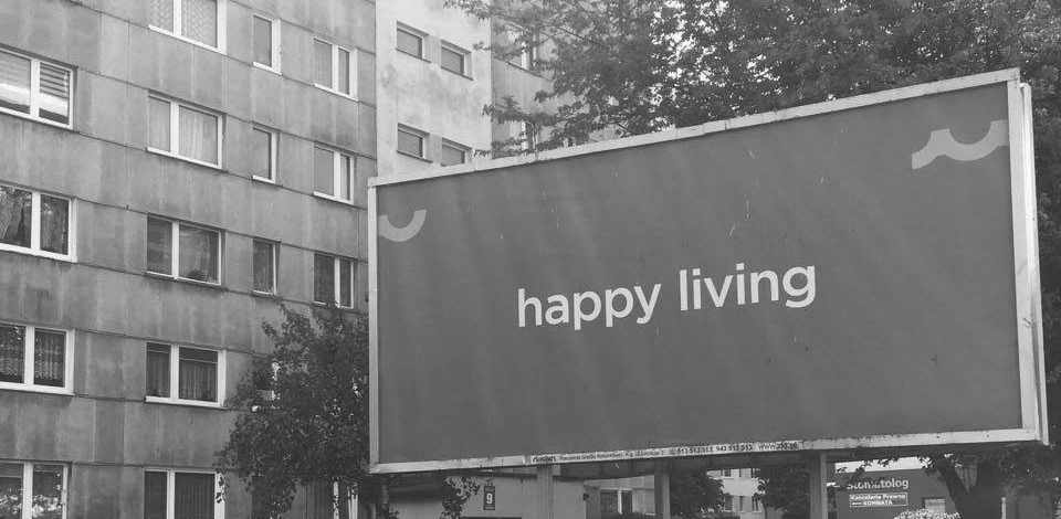 Plattenbau mit Werbeschild im Vordergrund mit Aufschrift "Happy Living". Grautöne.