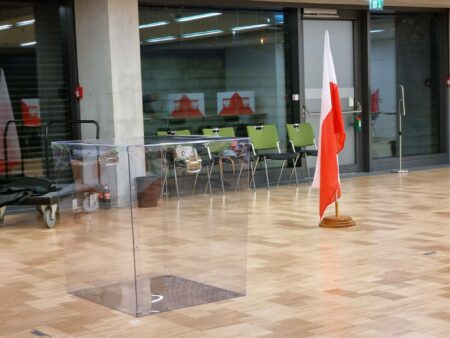 Das Wahlbüro in Darmstadt. Eine leere Urne steht im Vordergrund auf Parkettboden. Eine große polnische Flagge steht im Raum, im Hintergrund leere Stühle vor einer Glaswand. Foto: Antoni Stelter